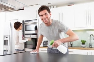 Man cleaning kitchen countertop with spray bottle, woman organizing kitchen appliances in modern remodeled kitchen, emphasizing the importance of professional plumbing in kitchen renovations.
