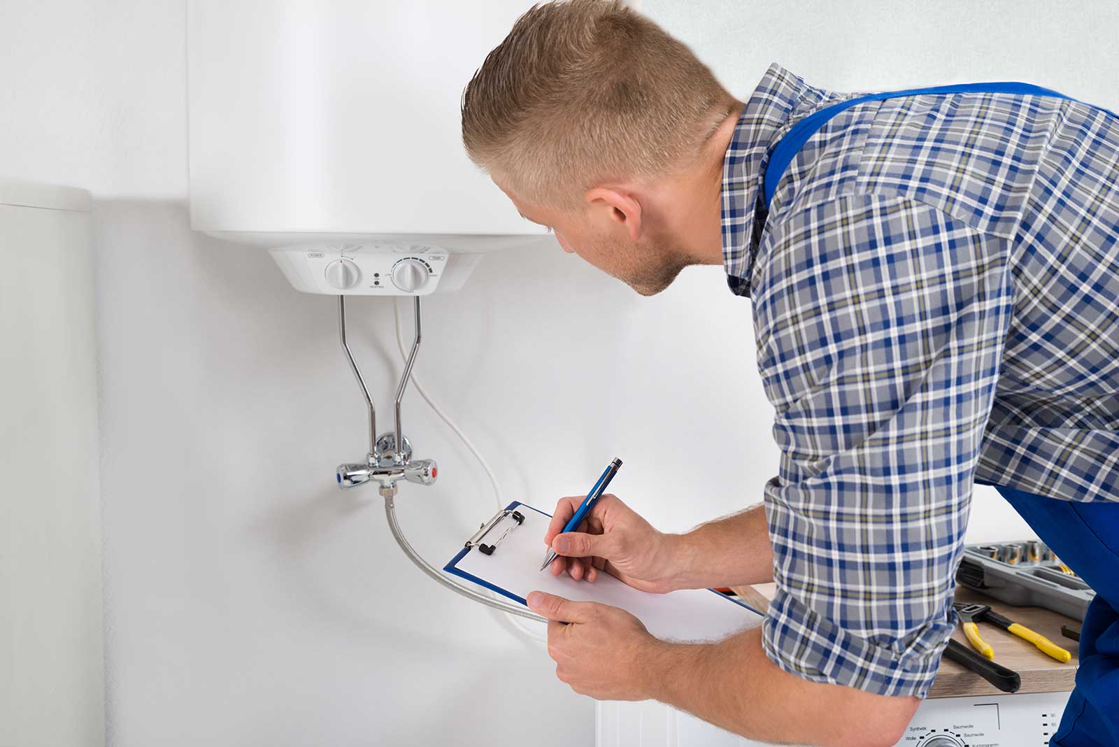 Technician servicing a water heater at home, wearing a plaid shirt and blue overalls, with tools on a surface.