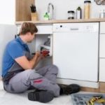 Man fixing a dishwasher in a modern kitchen, showcasing tools and household items; Allore's Plumbing Services LLC.