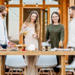 Four friends gather outdoors at a wooden table enjoying food and drinks, representing a relaxed social celebration.