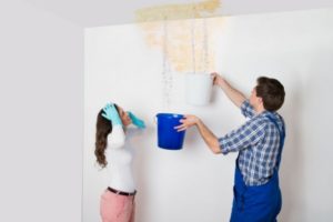 Man and woman managing a water leak, with buckets collecting dripping water from a ceiling, illustrating the importance of addressing plumbing issues to prevent water waste and save on bills.