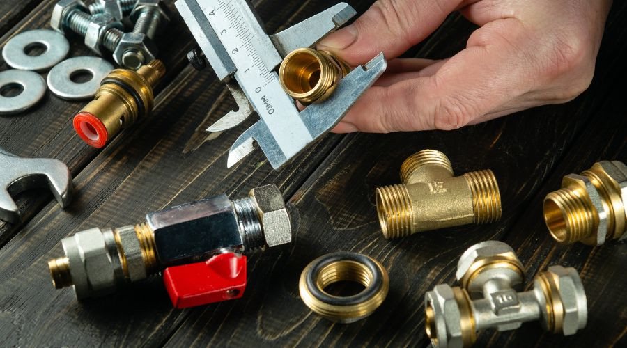 Hand holding a caliper measuring brass plumbing fittings and tools at Allore's Plumbing Services LLC workspace.