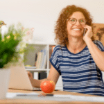 Woman with curly hair and glasses in home office on phone, representing Allore's Plumbing Services.