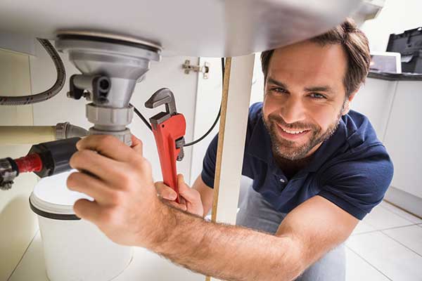 Plumber kneeling under kitchen sink, using adjustable wrench with visible plumbing pipes for home maintenance.