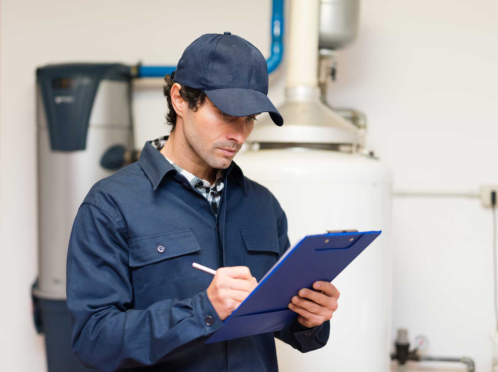 Plumber in uniform holding clipboard in work setting with plumbing equipment, focusing on maintenance tasks.